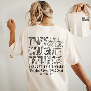 May include: Cream-colored t-shirt with black text and graphics. The back of the shirt reads "THEY CAUGHT FEELINGS" with a ring box and disco ball graphic. The text "I CAUGHT THIS T-SHIRT the Jackson Wedding 10.05.25" is also printed.
