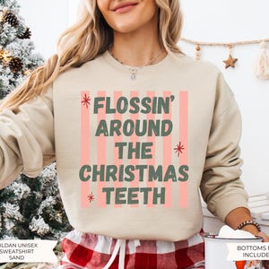 K&ouml;nnte beinhalten: Ein sandfarbenes Sweatshirt mit Rundhalsausschnitt und einem Grafikdruck aus vertikalen rosa Streifen sowie dem dunkelgr&uuml;nen Text FLOSSIN' AROUND THE CHRISTMAS TEETH, akzentuiert durch kleine rote Sternsymbole. Das Kleidungsst&uuml;ck hat lange &Auml;rmel.