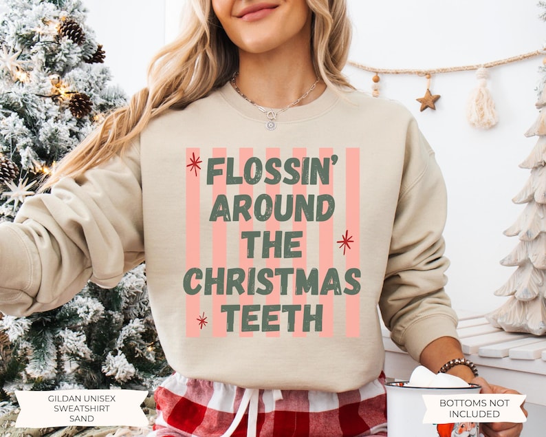 K&ouml;nnte beinhalten: Ein sandfarbenes Sweatshirt mit Rundhalsausschnitt und einem Grafikdruck aus vertikalen rosa Streifen sowie dem dunkelgr&uuml;nen Text FLOSSIN' AROUND THE CHRISTMAS TEETH, akzentuiert durch kleine rote Sternsymbole. Das Kleidungsst&uuml;ck hat lange &Auml;rmel.