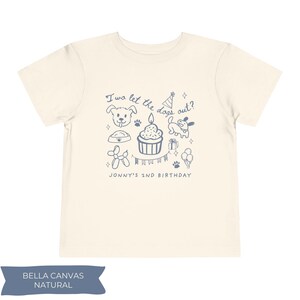 以下が含まれることがあります： クリーム色の子供用Tシャツ。青色のグラフィックには犬のイラスト、ろうそく付きのカップケーキ、そして「I wo let the dogs out?」と「JONNY'S 2ND BIRTHDAY」の文字が含まれています。