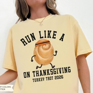 May include: A butter yellow t-shirt featuring a graphic of a running dinner roll wearing a headband. The black text reads RUN LIKE A ON THANKSGIVING TURKEY TROT 2026. The illustration shows the roll with stick arms and legs in a running pose.