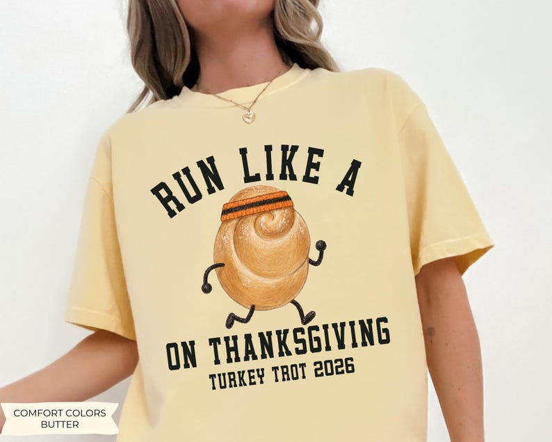 May include: A butter yellow t-shirt featuring a graphic of a running dinner roll wearing a headband. The black text reads RUN LIKE A ON THANKSGIVING TURKEY TROT 2026. The illustration shows the roll with stick arms and legs in a running pose.