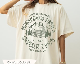 May include: A white t-shirt with a green graphic of a cabin in the mountains. The graphic says "Mountain Views Cate's Bachelorette Est. 2025 Before I Do's Blue Ridge, GA".