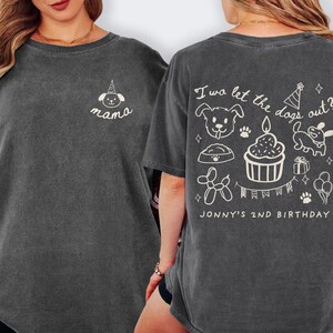 以下が含まれることがあります： クルーネックの濃いグレーのTシャツ。前面には、パーティーハットをかぶった犬の小さな白いグラフィックと「mama」の文字があります。背面には「I wa let the dogs out?」というフレーズと犬をテーマにしたイラスト、「JONNY'S 2ND BIRTHDAY」があります。