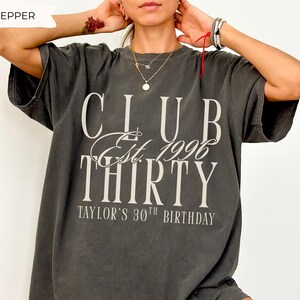 May include: A charcoal gray short-sleeve t-shirt featuring the white text CLUB Est. 1996 THIRTY and TAYLOR'S 30TH BIRTHDAY printed on the front. The shirt has a crew neckline and a relaxed fit, with the word PEPPER displayed in the top left corner.