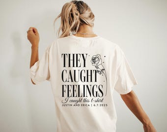 May include: Cream-colored t-shirt with the text "THEY CAUGHT FEELINGS" in a bold, black font. Below, it reads "I caught this t-shirt" followed by names and a date. A black floral design is on the right.