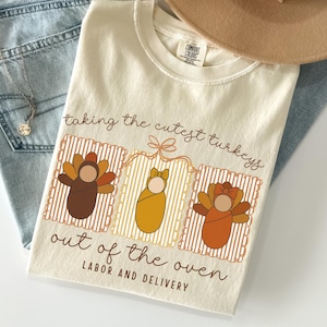 May include: Cream-colored t-shirt with a Thanksgiving-themed graphic. The design features three baby turkeys with the text "taking the cutest turkeys out of the oven LABOR AND DELIVERY."
