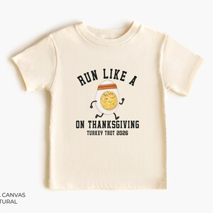 May include: A natural-colored short-sleeve t-shirt featuring a graphic of a running egg wearing an orange headband. The text reads RUN LIKE A [EGG] ON THANKSGIVING TURKEY TROT 2026 in black block lettering. The shirt is labeled BELLA CANVAS NATURAL.