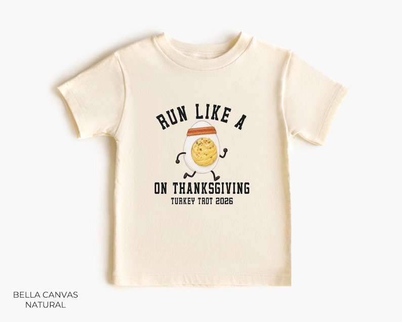 May include: A natural-colored short-sleeve t-shirt featuring a graphic of a running egg wearing an orange headband. The text reads RUN LIKE A [EGG] ON THANKSGIVING TURKEY TROT 2026 in black block lettering. The shirt is labeled BELLA CANVAS NATURAL.