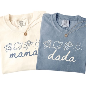 May include: A cream t-shirt with blue space-themed graphics and the word mama, and a blue t-shirt with white space-themed graphics and the word dada. Both shirts feature a rocket, planet, astronaut, and sun icon, with Comfort Colors labels.