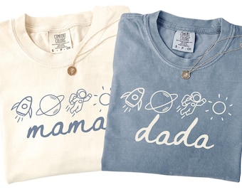 Puede incluir: Una camiseta color crema con gráficos espaciales azules y la palabra mama, y una camiseta azul con gráficos espaciales blancos y la palabra dada. Ambas camisetas presentan un cohete, un planeta, un astronauta y un icono de sol.
