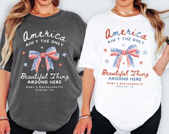 May include: Two t-shirts, one dark gray and one white, featuring the text "America Ain't The Only Beautiful Thing Around Here." The design includes a bow with red, white, and blue stripes and stars, perfect for a bachelorette party in Austin, TX.