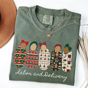 Peut inclure: T-shirt vert olive avec un motif de cinq b&eacute;b&eacute;s emmaillot&eacute;s, chacun avec un motif et un n&oelig;ud diff&eacute;rents. Le texte "Labor and Delivery" est brod&eacute; en dessous. Un collier en or est drap&eacute; sur le t-shirt.