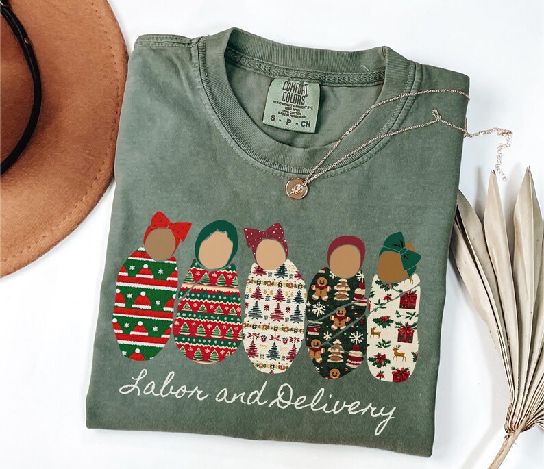 Peut inclure: T-shirt vert olive avec un motif de cinq b&eacute;b&eacute;s emmaillot&eacute;s, chacun avec un motif et un n&oelig;ud diff&eacute;rents. Le texte "Labor and Delivery" est brod&eacute; en dessous. Un collier en or est drap&eacute; sur le t-shirt.