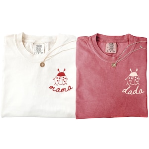 May include: Two t-shirts, one cream and one dusty rose, each with a ladybug graphic and text. The cream shirt says "mama" in red script, and the rose shirt says "dada" in white script. Both shirts have a Comfort Colors tag and a gold pendant necklace.