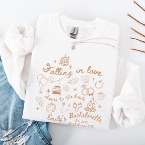 May include: A white crewneck sweatshirt with a gold graphic design. The design includes the text "Falling in love, cheers to the bride, Emily's Bachelorette, Gatlinburg, TN." and various illustrations such as a disco ball, balloons, and a campfire. A silver necklace is draped across the sweatshirt.
