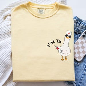 May include: A pale yellow t-shirt with a cartoon goose wearing sunglasses and a nurse's hat, holding a syringe. The text "Stick 'em" is printed on the shirt. The shirt is folded on a white surface with a pair of blue jeans and a phone.