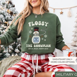 May include: Military green sweatshirt with the text "FLOSSY THE SNOWMAN" and a snowman graphic. The sweatshirt also has the text "HE'S A JOLLY, HAPPY ROLL". The image also shows red and white plaid pajama pants.