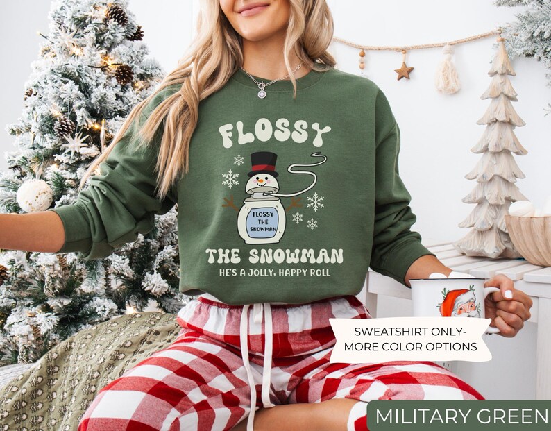 May include: Military green sweatshirt with the text "FLOSSY THE SNOWMAN" and a snowman graphic. The sweatshirt also has the text "HE'S A JOLLY, HAPPY ROLL". The image also shows red and white plaid pajama pants.