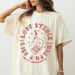 May include: Cream-colored t-shirt with a red graphic. The graphic reads "LOVE STRUCK LIKE A MATCH" in a circular design, with a match and matchbox illustration. Text includes "EST. 2026" and "EMILY'S BACHELORETTE".