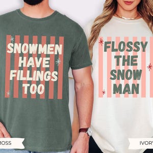 K&ouml;nnte beinhalten: Ein moosgr&uuml;nes T-Shirt mit dem Text SNOWMEN HAVE FILLINGS TOO und ein elfenbeinfarbenes T-Shirt mit dem Text FLOSSY THE SNOW MAN. Beide T-Shirts zeigen einen Hintergrund aus vertikalen korallenfarbenen Streifen und kleinen Sternen im Used-Look.