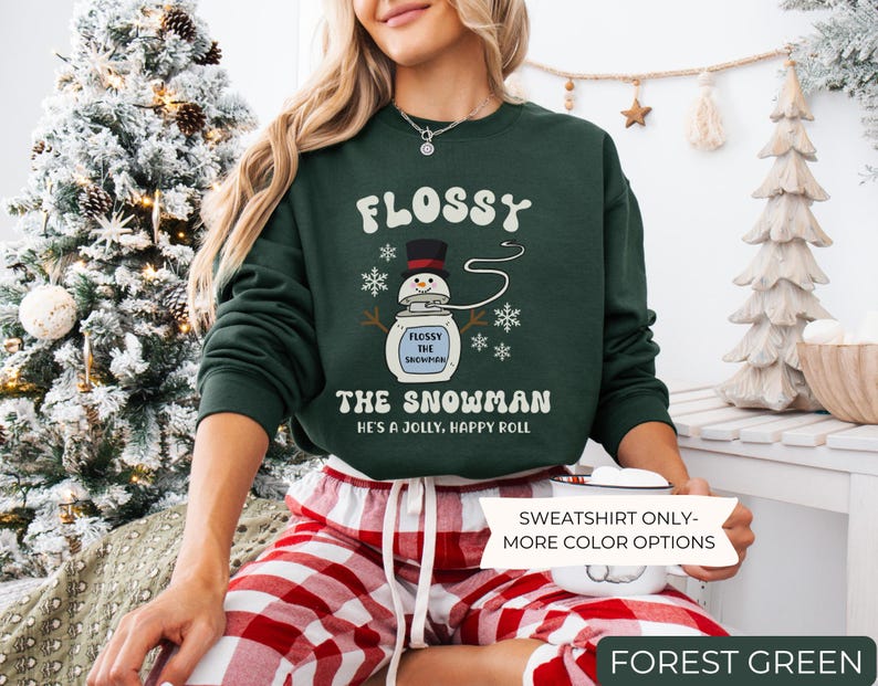 May include: A forest green sweatshirt with the text "FLOSSY THE SNOWMAN" and a cartoon snowman design. The sweatshirt also has the text "HE'S A JOLLY, HAPPY ROLL". The image also includes the text "SWEATSHIRT ONLY - MORE COLOR OPTIONS" and "FOREST GREEN".