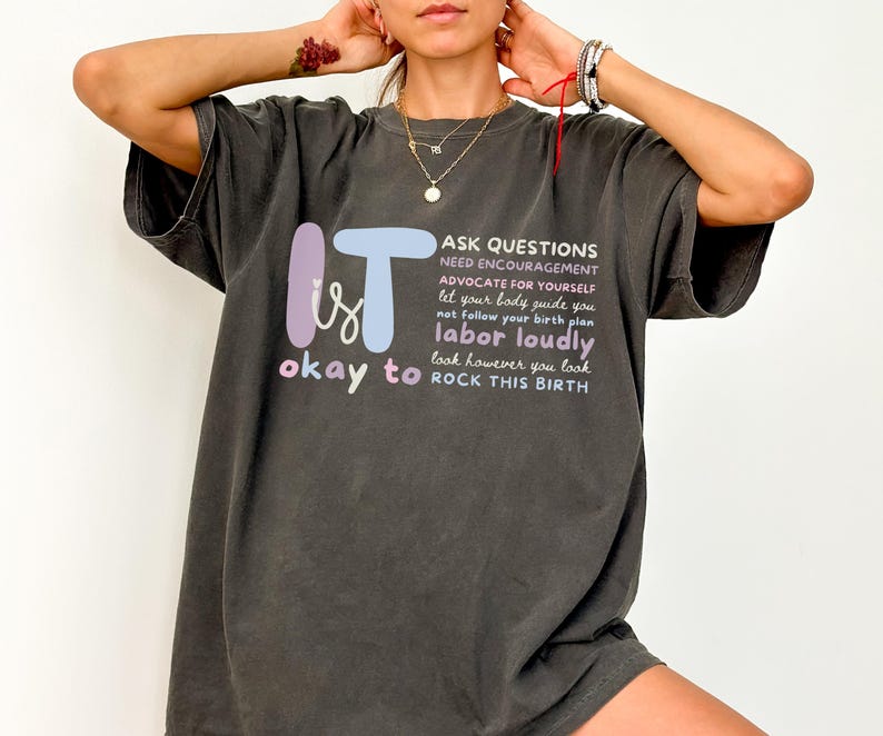 May include: Dark gray t-shirt with the words "IT is okay to" in a colorful design. The shirt also features the phrases "ASK QUESTIONS, labor loudly, ROCK THIS BIRTH" in white text. A casual, comfortable style.