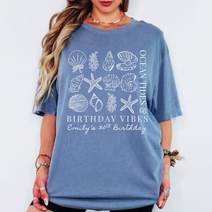 May include: A blue t-shirt with a white seashell and starfish design. The shirt has the text "OCEAN TIDES & BIRTHDAY VIBES" and "Emily's 30th Birthday". The shirt is a casual, comfortable style.