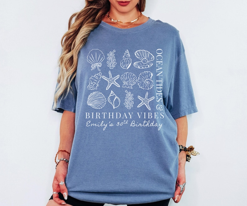 May include: A blue t-shirt with a white seashell and starfish design. The shirt has the text "OCEAN TIDES & BIRTHDAY VIBES" and "Emily's 30th Birthday". The shirt is a casual, comfortable style.