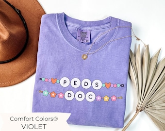 May include: Purple t-shirt with the text "PEDS DOC" in colourful letters and beads. The shirt is from Comfort Colors.