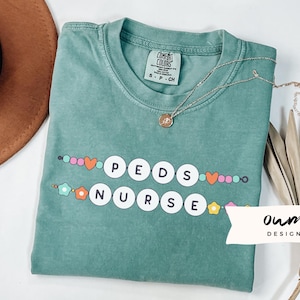 May include: A teal blue t-shirt with the words "PEDS NURSE" in white letters surrounded by colorful beads and flowers. The shirt is made of a soft, comfortable material and is perfect for everyday wear.