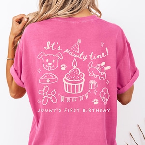 May include: A magenta t-shirt with white graphics and text. The design includes a dog, a cupcake with a candle, a party hat, a dog bowl, a balloon dog, a gift, paw prints, and balloons. The text reads "It's pawty time!" and "Jonny's First Birthday".