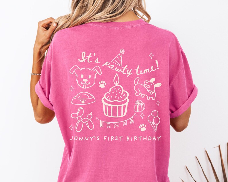 May include: A magenta t-shirt with white graphics and text. The design includes a dog, a cupcake with a candle, a party hat, a dog bowl, a balloon dog, a gift, paw prints, and balloons. The text reads "It's pawty time!" and "Jonny's First Birthday".