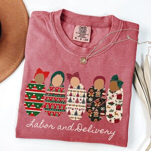 Peut inclure: Un t-shirt rose fan&eacute; avec les mots "Labor and Delivery" en &eacute;criture blanche. Le t-shirt pr&eacute;sente un graphisme de cinq b&eacute;b&eacute;s emmaillot&eacute;s avec diff&eacute;rentes couvertures &agrave; motifs et des n&oelig;uds. Un collier en argent avec un petit pendentif est drap&eacute; sur le t-shirt.