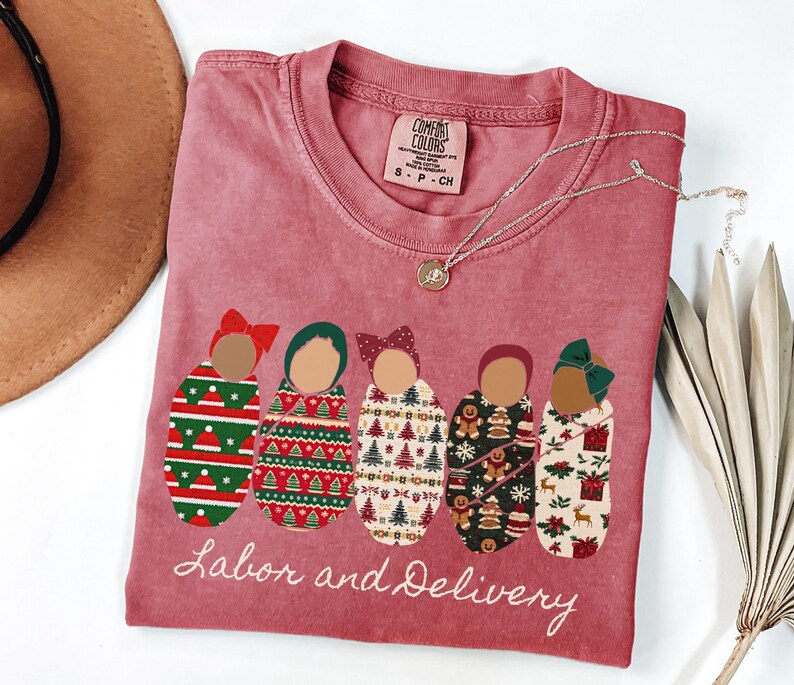 Peut inclure: Un t-shirt rose fan&eacute; avec les mots "Labor and Delivery" en &eacute;criture blanche. Le t-shirt pr&eacute;sente un graphisme de cinq b&eacute;b&eacute;s emmaillot&eacute;s avec diff&eacute;rentes couvertures &agrave; motifs et des n&oelig;uds. Un collier en argent avec un petit pendentif est drap&eacute; sur le t-shirt.