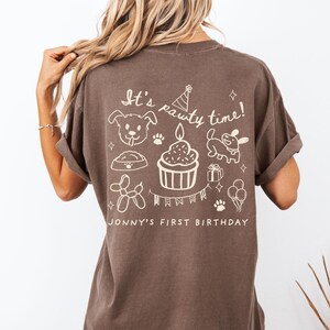 May include: Brown t-shirt with white graphic text and illustrations. The text reads "It's pawty time!" with images of a dog, paw prints, a cupcake, and balloons. Below the text is "Jonny's First Birthday".
