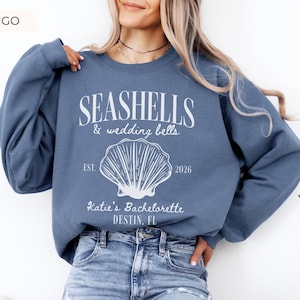 May include: Indigo blue sweatshirt with white text that reads "Seashells & wedding bells" with a seashell graphic. Below the graphic, the text reads "Katie's Bachelorette DESTIN, FL" and "EST. 2026".
