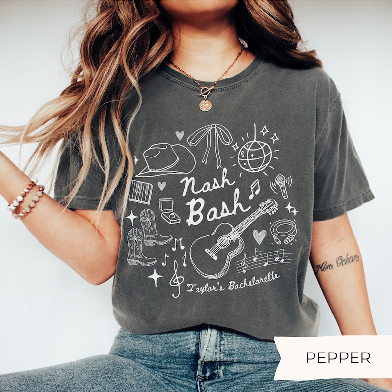 May include: A dark grey t-shirt with a white graphic design featuring a guitar, cowboy boots, a microphone, a disco ball, and the text "Nash Bash Taylor's Bachelorette".