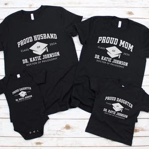 May include: Black t-shirts with white text and a graduation cap graphic. The shirts say "Proud Husband", "Proud Mom", and "Proud Daughter" with the year 2024 and the name "Dr. Katie Johnson, Doctor of Philosophy".