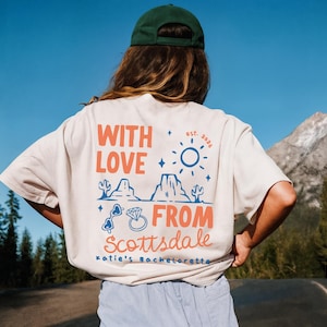 May include: Cream-colored t-shirt with a graphic design on the back. The design includes the words "WITH LOVE FROM Scottsdale" in orange and blue, along with illustrations of cacti, mountains, and a ring. The text "Katie's Bachelorette" is also included.