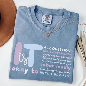 May include: A blue Comfort Colors t-shirt with the text "It is okay to labor loudly, rock this birth." The shirt has a crew neck and short sleeves. A gold necklace with a pendant is draped across the shirt.