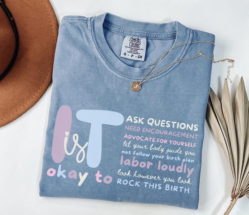 May include: A blue Comfort Colors t-shirt with the text "It is okay to labor loudly, rock this birth." The shirt has a crew neck and short sleeves. A gold necklace with a pendant is draped across the shirt.