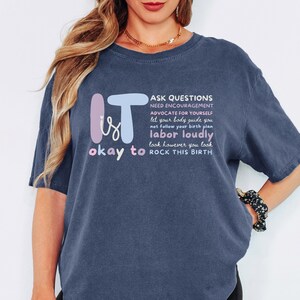 May include: A navy blue t-shirt with the words "IT is okay to" in a whimsical font, and a list of phrases about childbirth. The shirt has short sleeves and a relaxed fit.