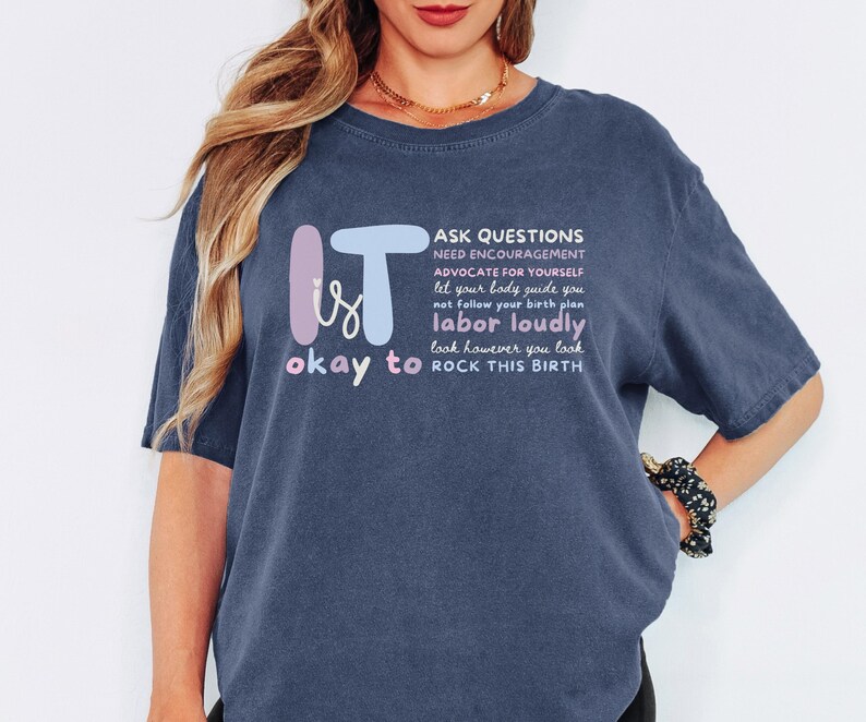 May include: A navy blue t-shirt with the words "IT is okay to" in a whimsical font, and a list of phrases about childbirth. The shirt has short sleeves and a relaxed fit.