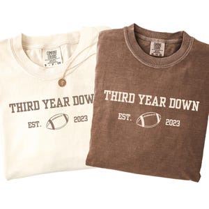 May include: Two t-shirts, one cream and one brown, with the text "THIRD YEAR DOWN" and a football graphic. Below the text is "EST. 2023". The cream shirt has a gold necklace. Both shirts are Comfort Colors brand.