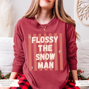 K&ouml;nnte beinhalten: Ein karmesinrotes Langarm-Sweatshirt mit Rundhalsausschnitt und einem Grafikdruck mit vertikalen Streifen in einem helleren Rotton. Der Text FLOSSY THE SNOW MAN ist in wei&szlig;en Blockbuchstaben gehalten und mit kleinen hellblauen Sternen verziert.