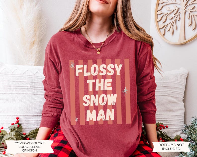 K&ouml;nnte beinhalten: Ein karmesinrotes Langarm-Sweatshirt mit Rundhalsausschnitt und einem Grafikdruck mit vertikalen Streifen in einem helleren Rotton. Der Text FLOSSY THE SNOW MAN ist in wei&szlig;en Blockbuchstaben gehalten und mit kleinen hellblauen Sternen verziert.