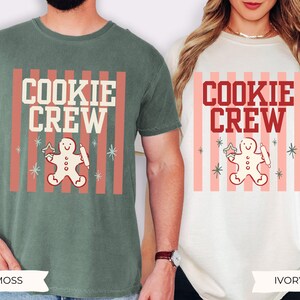 May include: A moss green t-shirt and an ivory t-shirt, each featuring a graphic print with vertical pink stripes, the text "COOKIE CREW" in bold lettering, and a central illustration of a gingerbread person holding a star and a rolling pin.