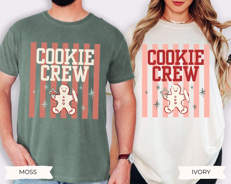May include: A moss green t-shirt and an ivory t-shirt, each featuring a graphic print with vertical pink stripes, the text "COOKIE CREW" in bold lettering, and a central illustration of a gingerbread person holding a star and a rolling pin.