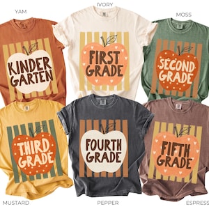 May include: Six short-sleeve t-shirts in yam, ivory, moss, mustard, pepper, and espresso colors. Each shirt features a central graphic of an apple with vertical stripes behind it and text reading Kindergarten, First Grade, Second Grade, Third Grade, Fourth Grade, or Fifth Grade.